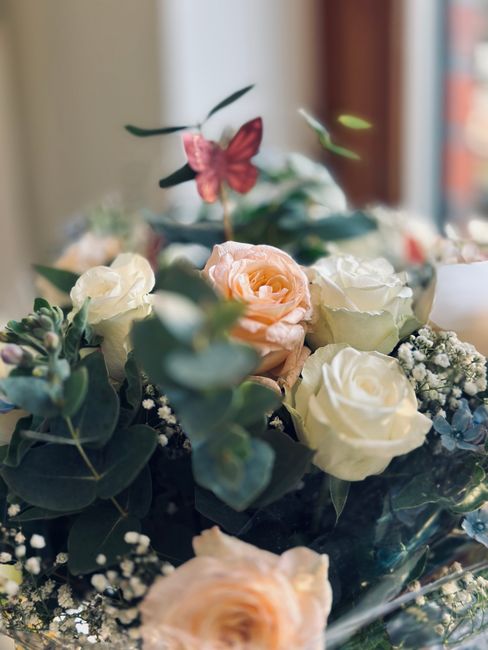 A close-up view of a floral arrangement featuring pale peach and cream roses, complemented by sprigs of white baby's breath and lush green eucalyptus leaves. The bouquet is set in a dark, glossy vase,